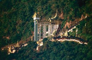 A Buddhist monastery is nestled into a lush green hillside with tiered buildings, ornate roofs, and a striking elevator tower.