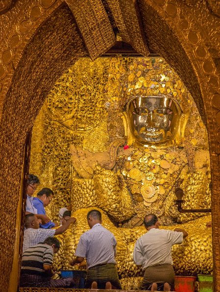 A group of men are performing a ritual in front of a massive, gold-covered Buddha statue inside a richly decorated temple.