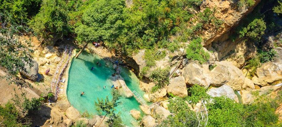 An aerial view captures a vibrant turquoise natural pool surrounded by lush greenery and rocky cliffs, with a small wooden bridge leading to the water where several people are swimming.