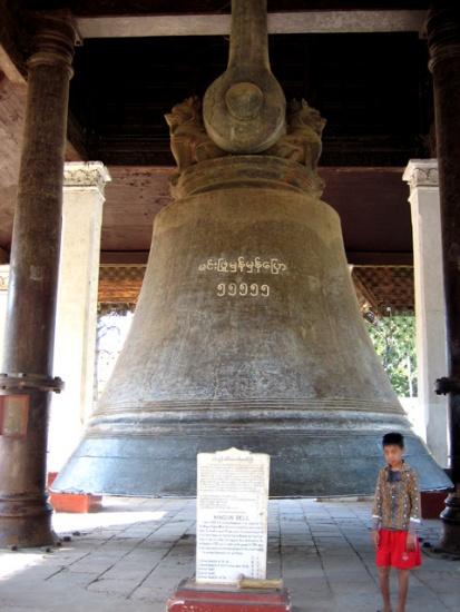 A young boy in a patterned shirt and red shorts stands next to a large bell inscribed with Burmese script.