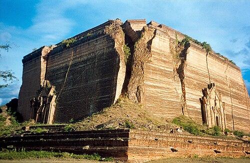 A colossal, unfinished brick stupa stands under a blue sky, showing the layered construction and dramatic erosion of its massive structure.