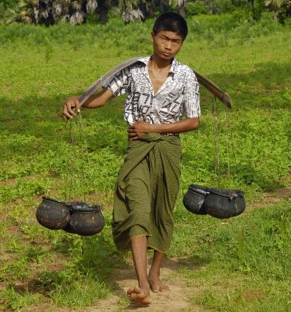 A young man walks through a grassy field with two pots hanging from a yoke across his shoulders.