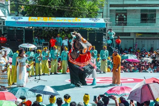 A group of performers in traditional attire are participating in a cultural event, possibly a parade or festival, with a large crowd watching.