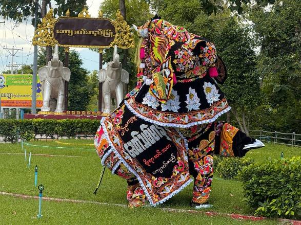 A dancer in an elaborate elephant costume performs outdoors in front of a decorative archway with elephant statues.
