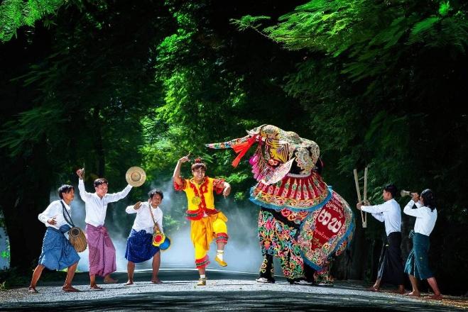 A group of performers in traditional clothing are dancing and playing instruments in front of a large, ornate elephant costume on a tree-lined path.