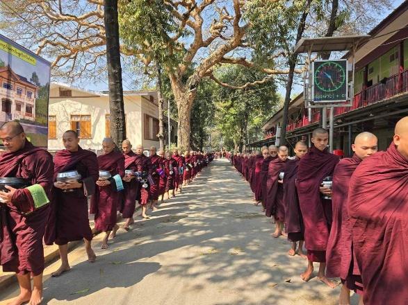 A long line of Buddhist monks in maroon robes walks down a street, holding alms bowls.