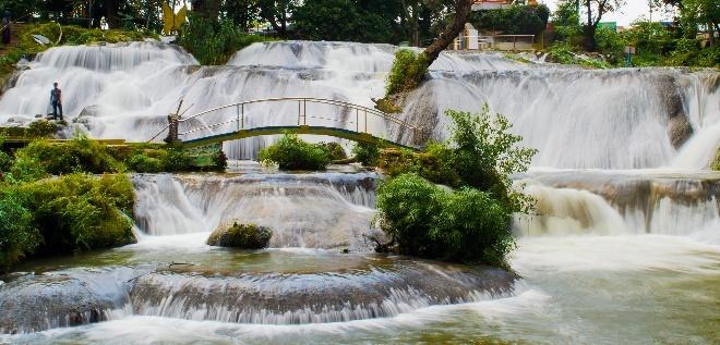 A picturesque cascade flows over tiered rocks, with a charming green bridge spanning the water and lush vegetation framing the scene.