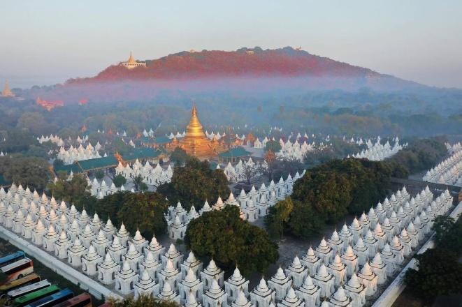 An aerial view shows a vast complex of white stupas surrounding a golden temple, with a mist-covered, red-hued mountain in the background.