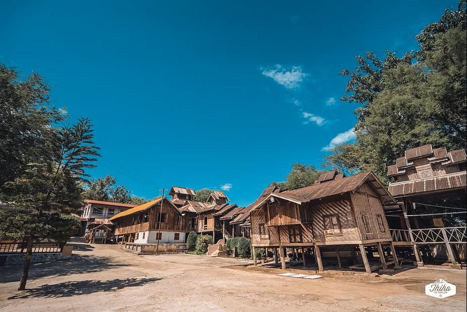 A village of stilted wooden houses with rustic roofs sits under a bright blue sky with scattered clouds.