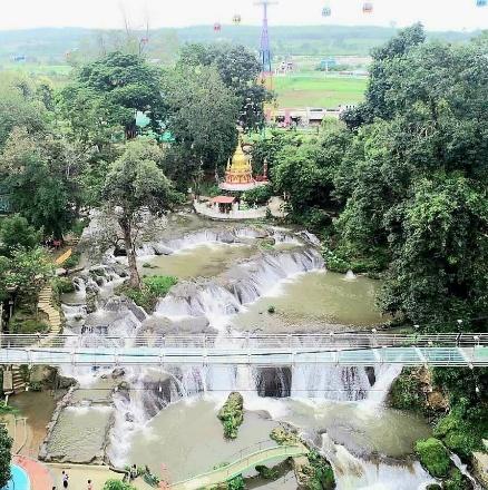 A scenic aerial view showcases a multi-tiered waterfall cascading through lush greenery, with a golden pagoda and a glass bridge adding cultural and architectural elements to the landscape.