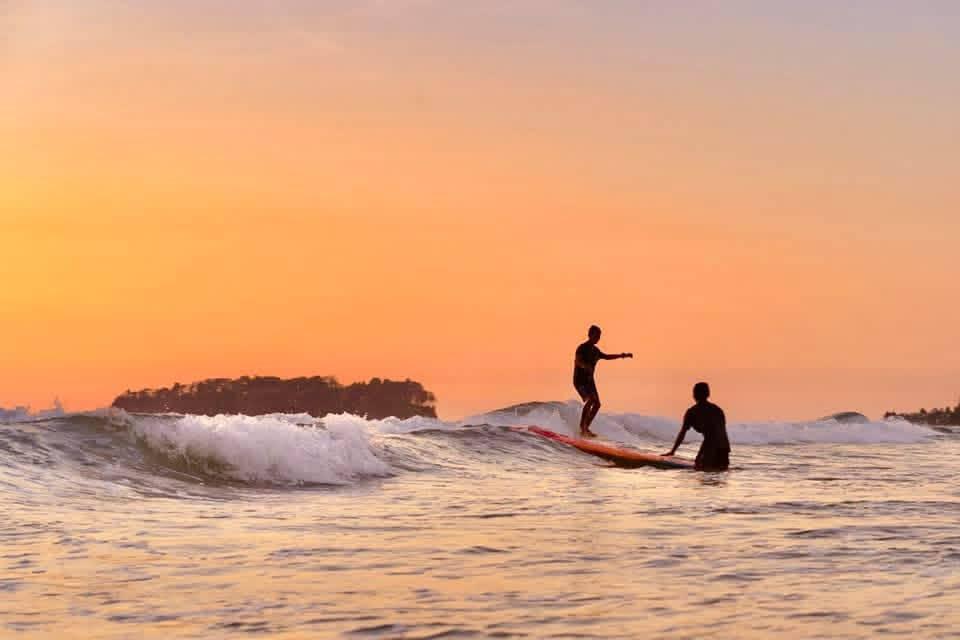 Two surfers ride waves against a vibrant orange sunset with a silhouetted island in the background.