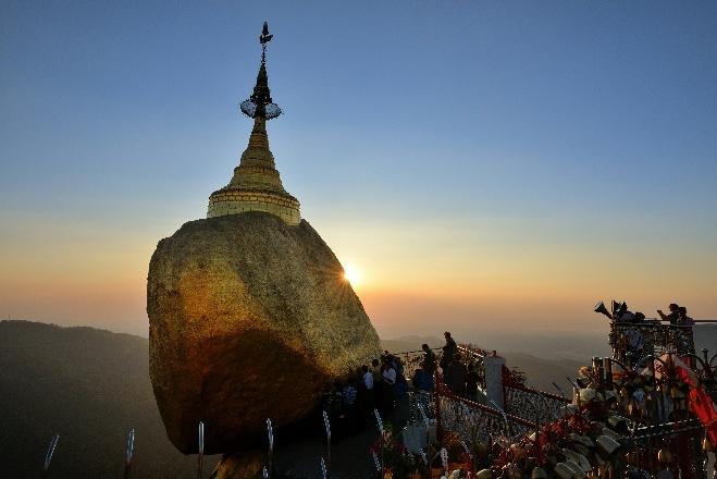 A golden Buddhist pagoda rests precariously atop a massive boulder against a sunrise sky.