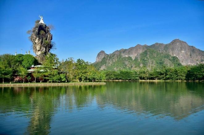 A white stupa sits atop a rocky outcrop surrounded by lush greenery and reflected in a calm lake, with a majestic mountain range in the background.
