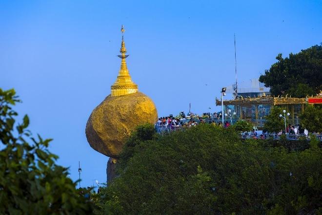 A large golden stupa crowns a boulder precariously balanced on a cliff face, with a temple complex and crowds of people visible on the hilltop.