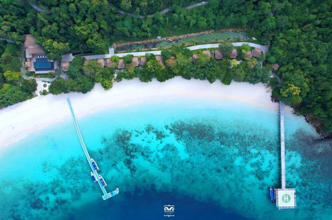 An aerial view shows a serene tropical resort with lush greenery, a white sand beach, crystal-clear turquoise waters, and piers extending into the sea.