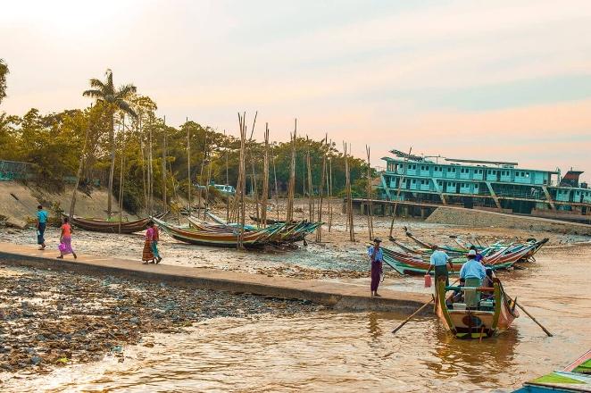 A group of colorful boats are moored on a rocky shore in front of lush trees and a large blue multi-story building.