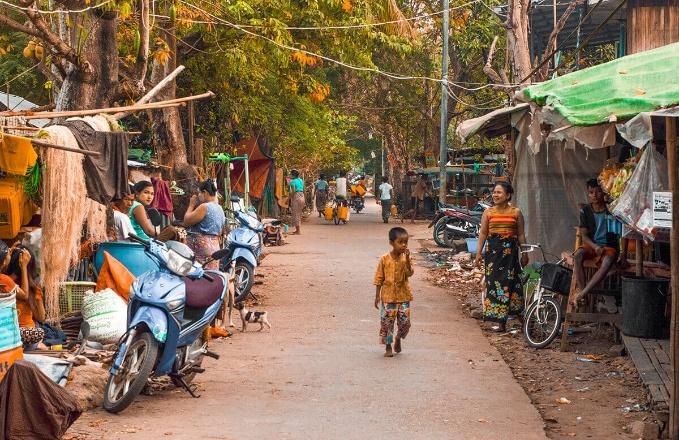 A young child walks down a street lined with shops and motorcycles, with people and colorful fabrics adding to the vibrant scene.